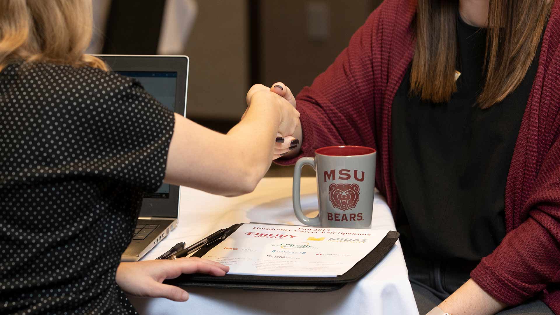 Two people shaking hands over a notepad and Missouri State coffee mug.