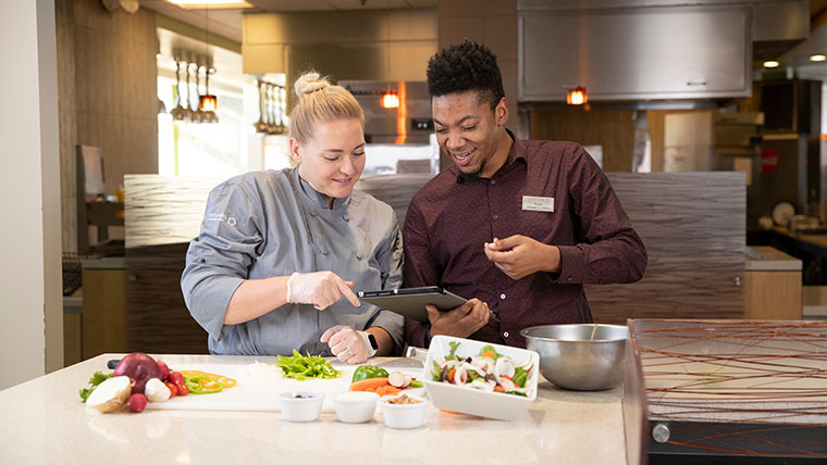 Hospitality alum uses a tablet computer while working in the kitchen for the Blair-Shannon dining hall. The table in front of them has veggies in bowls and on a cutting board.